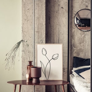 Puede incluir: Una mesa de centro ovalada de madera con una maceta marr&oacute;n, un jarr&oacute;n de vidrio marr&oacute;n con ramas secas y una escultura de mano de madera. Un dibujo en blanco y negro enmarcado de tulipes cuelga de la pared detr&aacute;s de la mesa. La mesa est&aacute; delante de una cama con s&aacute;banas blancas y una almohada azul oscuro.