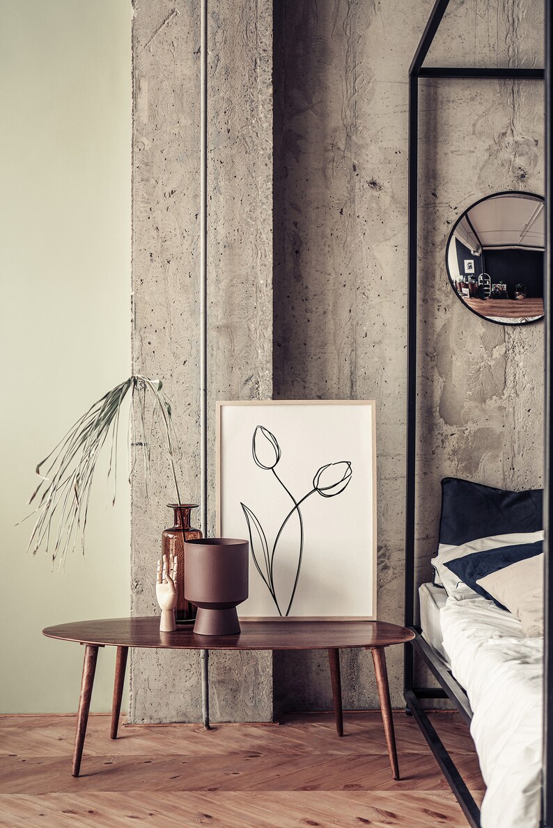 Puede incluir: Una mesa de centro ovalada de madera con una maceta marr&oacute;n, un jarr&oacute;n de vidrio marr&oacute;n con ramas secas y una escultura de mano de madera. Un dibujo en blanco y negro enmarcado de tulipes cuelga de la pared detr&aacute;s de la mesa. La mesa est&aacute; delante de una cama con s&aacute;banas blancas y una almohada azul oscuro.