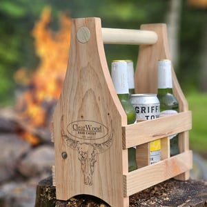 May include: A wooden beer caddy with a handle, holding several bottles and a can. The caddy is made of light-coloured wood and features a bull skull design with the text "ClearWood Beer Caddy" on the side. The caddy is sitting on a tree stump.