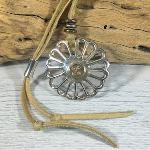 以下が含まれることがあります： 中央にカットアウトされた花のデザインと小さな真鍮製の矢印が付いた銀製のペンダント。ペンダントは茶色の革紐に取り付けられています。