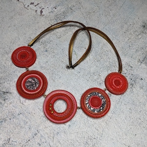 Puede incluir: Un collar rojo y dorado con cinco colgantes circulares. Los colgantes tienen diferentes patrones y diseños. El collar está hecho de cordón de cuero.