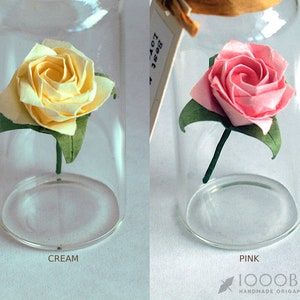 May include: Two origami roses in glass jars. The rose on the left is cream colored and the rose on the right is pink. Both roses have green leaves.