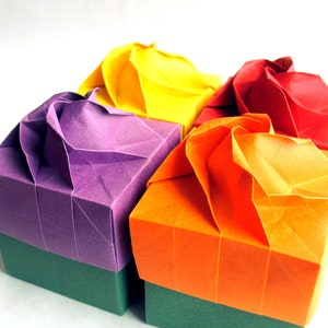 Puede incluir: Cuatro cajas de origami en diferentes colores: morado, naranja, amarillo y rojo. Las cajas están dobladas en una forma decorativa de flor.