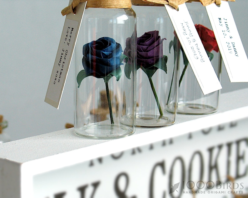 May include: Three glass bottles with cork tops, each containing a single origami rose. The roses are blue, purple, and red. Each bottle has a small tag attached with handwritten text. The text on the tags reads "Merry Christmas Love. Jinny xoxo", "Jinny & Danny May 10, 2019", and "Jinny & Danny May 10, 2019".