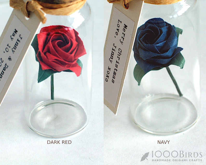 May include: Two glass jars with cork lids, each containing a single origami rose. The rose in the left jar is dark red, and the rose in the right jar is navy blue. The jars have handwritten tags attached to them.