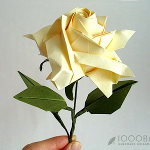 May include: A delicate origami rose with a cream-coloured flower and green leaves. The rose is handmade and appears to be crafted from paper.