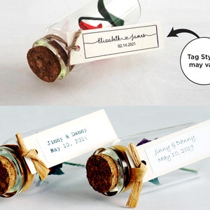 May include: Two glass bottles with cork stoppers and a small origami flower inside. Each bottle has a white tag tied around the neck with handwritten text. The top bottle's tag reads "Elizahth James 02.14.2021". The bottom bottle's tag reads "Jinny & Danny May 10, 2019". The text "Tag Styles may vary" is written in a circle with an arrow pointing to the bottles.