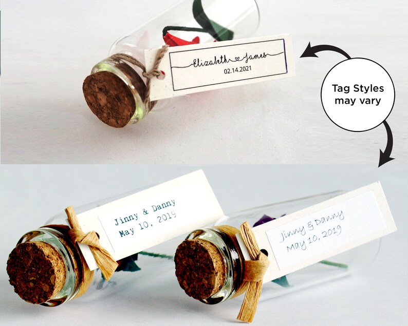 May include: Two glass bottles with cork stoppers and a small origami flower inside. Each bottle has a white tag tied around the neck with handwritten text. The top bottle's tag reads "Elizahth James 02.14.2021". The bottom bottle's tag reads "Jinny & Danny May 10, 2019". The text "Tag Styles may vary" is written in a circle with an arrow pointing to the bottles.