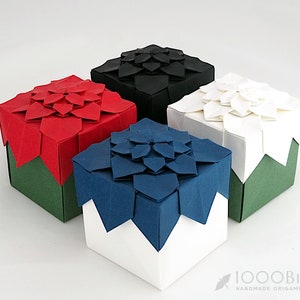 Może przedstawiać: Cztery pudełka origami z różnokolorowymi pokrywkami. Pudełka są wykonane z zielonego papieru z białymi podstawami. Pokrywki są czerwone, czarne, białe i niebieskie.