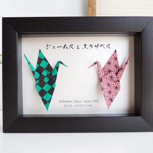 May include: A black shadow box frame containing two origami cranes on a white background. One crane has a green and black checkered pattern, the other a pink and black geometric pattern. Text reads: ジェームス & エリザベス, Akihabara, Tokyo - Japan 2026, JULY 29 - AUGUST 14, 2026.