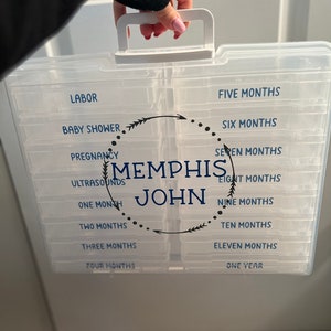 May include: Clear plastic storage box with a white handle, labeled with blue text. The box is divided into sections with labels such as "Labor," "Baby Shower," and "One Month." The name "Memphis John" is also displayed.