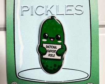 May include: A green enamel pin featuring a smiling pickle holding a sign that reads "Emotional Support Pickle." The pin is set against a light blue background with a jar illustration and the words "Pickles" in embossed letters. The pin is on a green card.