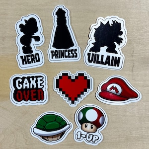 May include: Nine black and white stickers with various video game characters and phrases. The stickers include a hero, princess, villain, game over, heart, mushroom, and a turtle shell. The stickers are on a wood background.