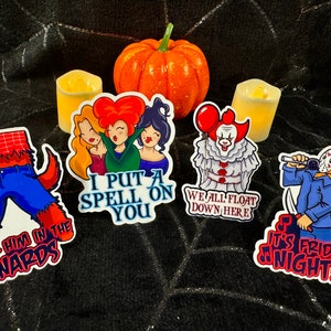 May include: A collection of Halloween-themed stickers featuring iconic characters and phrases. Designs include a werewolf, the Sanderson sisters, Pennywise the clown, and Jason Voorhees. The stickers have text like "Kick Him in the Nards" and "We All Float Down Here."