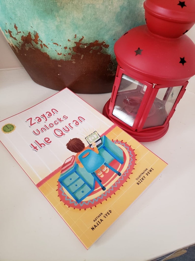 May include: A yellow and red children's book titled "Zayan Unlocks the Quran" with an illustration of a child sitting at a desk with a laptop. The book has the author's name "Naila Syed" and the illustrator's name "Rizky Dewi" printed on the back cover.