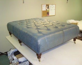 May include: A large, rectangular, tufted ottoman with a light blue fabric upholstery. The ottoman has four wooden legs with castors.