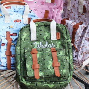 Könnte beinhalten: Eine Auswahl an bunten Rucksäcken mit verschiedenen Mustern, darunter Dinosaurier, Baufahrzeuge und Schmetterlinge. Jeder Rucksack hat braune Lederriemen und -griffe. Auf einem Rucksack steht der Name "Nikolas".