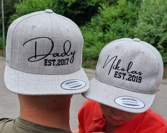 Op de afbeelding: Twee grijze baseballpetten met zwarte geborduurde tekst. De eerste pet zegt "Daddy Est. 2017" en de tweede pet zegt "Nikolas Est. 2019".