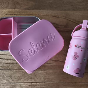 Peut inclure: Une boîte à lunch rose avec le nom "Selena" sur le couvercle, à côté d'une gourde rose assortie. La gourde a une poignée et est décorée de papillons et du nom "Selena".