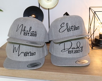 Op de afbeelding: Vier grijze baseballpetten met zwarte geborduurde tekst. De petten hebben de tekst "Mama est. 2017", "Daddy est. 2017", "Elias est. 2017" en "Nikolas est. 2019".