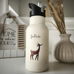 Può includere: Una borraccia bianca con tappo nero e illustrazione di cervo marrone. Il nome "Julia" è scritto in corsivo. Un vaso decorativo e un cartello incorniciato con la scritta "HOME is where YOUR is" sono sullo sfondo.