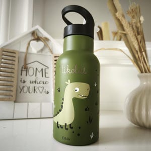 Può includere: Una borraccia verde con manico nero e grafica di dinosauro. Sulla bottiglia è stampato il nome "Nikolas". Sullo sfondo un'incorniciatura decorativa bianca a forma di casa con le parole "HOME is where YOUR ♡ is".
