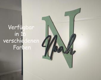 Op de afbeelding: Een houten wanddecoratie met de naam "Noah" in een cursief lettertype, over een grote groene letter "N". De tekst "Verfügbar in 16 verschiedenen Farben" is zichtbaar in de afbeelding.