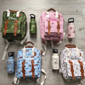 Könnte beinhalten: Eine Sammlung personalisierter Rucksäcke und Wasserflaschen. Die Rucksäcke zeigen Namen und verschiedene Muster, darunter Dinosaurier, Schmetterlinge, Lastwagen und Hirsche. Die Wasserflaschen haben passende Designs und Namen. Die Taschen haben braune Lederriemen und weiße Griffe.