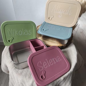 Può includere: Una collezione di portapranzo personalizzati in vari colori, tra cui verde, beige, blu e rosa. Ogni scatola presenta un coperchio con un nome inciso. Una scatola è aperta, rivelando un interno in acciaio inossidabile con un divisore.