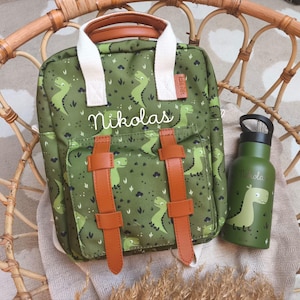 Könnte beinhalten: Grünes Rucksack- und Trinkflaschen-Set mit Dinosaurier-Muster. Der Rucksack hat braune Riemen und einen Griff, mit dem Namen "Nikolas" auf der Vorderseite. Die Trinkflasche trägt ebenfalls den Namen "Nikolas" und eine Dinosaurier-Grafik.