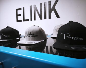 Op de afbeelding: Een collectie baseballpetten met platte klep in zwart en grijs, elk geborduurd met een naam en het oprichtingsjaar. De petten worden tentoongesteld op een reflecterend oppervlak met het woord "ELINIK" op de muur.