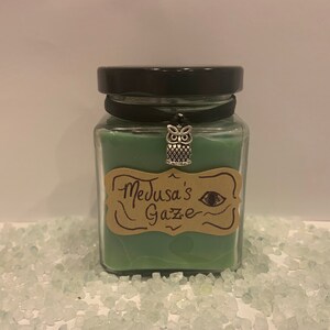 Puede incluir: Una vela verde en un tarro de vidrio cuadrado con tapa negra. La vela tiene una etiqueta marrón que dice "Medusa's Gaze" y un amuleto de búho plateado que cuelga de la tapa.