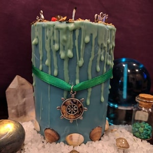 May include: A teal blue candle with green wax drippings and a silver ship's wheel charm. The candle is surrounded by seashells and white salt.