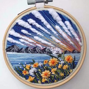 May include: Embroidered hoop art depicting a landscape with a blue sky, white clouds, mountains, a lake, and yellow flowers. The embroidery is done in a variety of colors and stitches.
