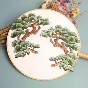 May include: A white fabric fan with a gold trim and three embroidered pine trees in shades of green and brown.