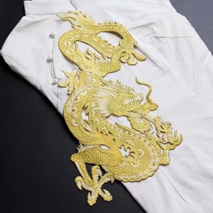 May include: A gold embroidered dragon applique patch. The dragon is curled up with its head facing forward and its tail curled around its body. The patch is detailed with intricate scales and a textured design.