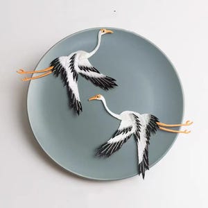 May include: Two embroidered crane patches featuring white bodies with black wing tips and orange beaks and legs. The birds are depicted in a flying position with wings spread wide, showcasing detailed stitching and contrasting colours on a round plate.