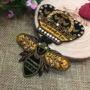 May include: A black velvet crown with gold and white embellishments and a black and gold embroidered bee with yellow and white beads.