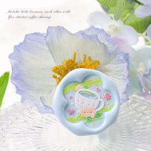 Può includere: Un sigillo di cera blu con un design di una tazza da tè e un piattino bianchi. La tazza da tè ha un bordo verde e un fiore rosa sul lato. Il sigillo è circondato da foglie verdi e fiori rosa.
