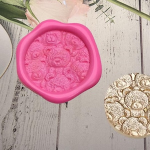 Puede incluir: Un molde rosa en forma de corazón lleno de diseños de osos de peluche, junto a un molde circular dorado con el mismo patrón de osos. Los moldes están sobre una superficie de madera clara, con acentos florales en el fondo. Estos moldes son probablemente para manualidades o repostería.