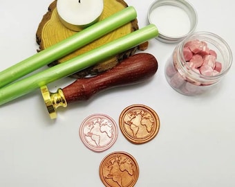 May include: A set of wax seal supplies including two green wax sticks, a wooden handle with a gold metal seal head, a jar of pink wax beads, and three wax seals with a world map design. Two of the seals are pink and one is copper.
