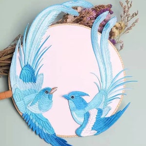 May include: Two blue embroidered bird appliques with white accents on a white background. The birds are facing each other and have long, flowing tails.