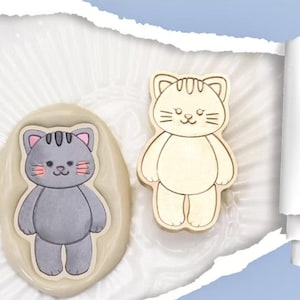May include: Two cat-shaped cookie cutters, one is a light brown color and the other is a gray color. Both cutters have a simple design with a rounded body, ears, and a face with eyes, a nose, and a mouth.