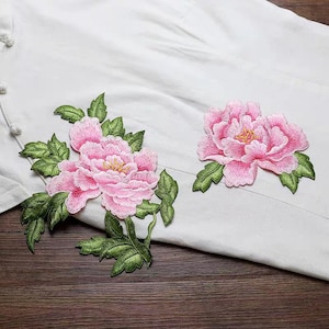 Op de afbeelding: Twee roze geborduurde bloemenapplicaties met groene bladeren. De bloemen zijn in een pioenroos-stijl.