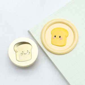 May include: A brass stamp featuring a raised design of a smiling cartoon bread slice. The stamp is round and has a smooth, polished surface. The bread is yellow and has a pink blush on its cheeks.
