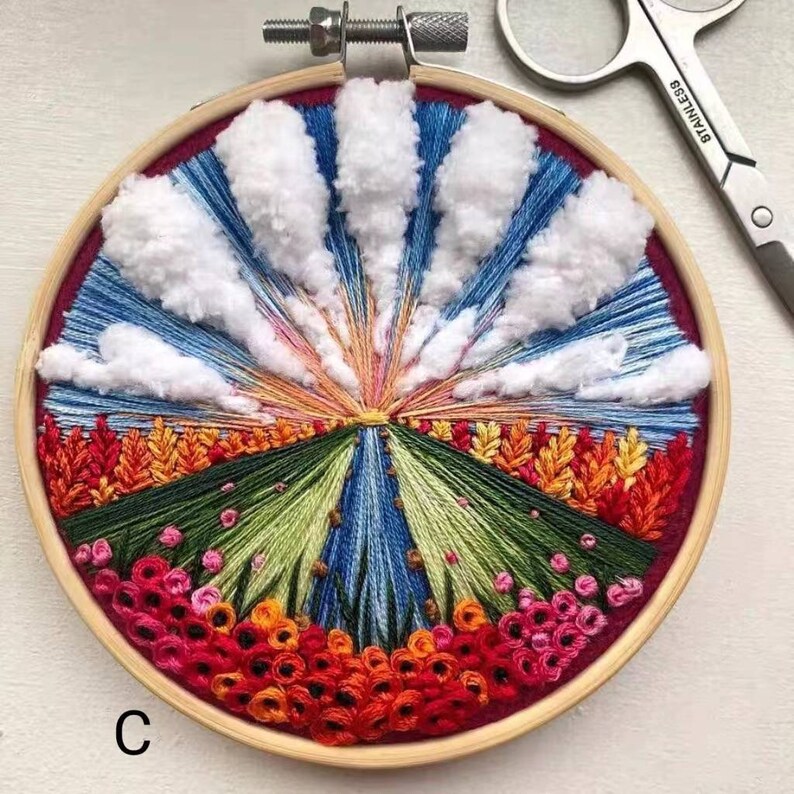 May include: A circular embroidery hoop with a colorful landscape scene. The scene features a blue sky with white fluffy clouds, a green field with orange flowers, and a bright orange sunset. The embroidery is framed in a wooden hoop with a red felt backing.