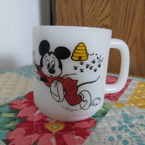 Pode incluir: Caneca de cer&acirc;mica branca com uma ilustra&ccedil;&atilde;o em preto e branco do Mickey Mouse fugindo de uma colmeia amarela e abelhas. A caneca tem uma al&ccedil;a preta.