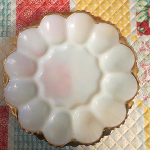 May include: A white, scalloped egg serving dish with a gold rim. The dish has 12 individual egg compartments and a central well. The dish is on a colorful patchwork quilt.