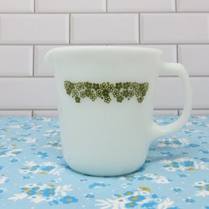 May include: White milk glass pitcher with a green floral design around the top. The pitcher has a handle and a rounded shape.