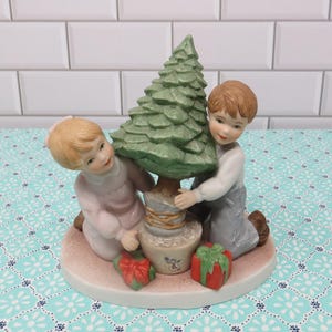 May include: A porcelain figurine of two children holding a small Christmas tree. The girl is wearing a pink dress and the boy is wearing blue jeans and a white shirt. There are two red wrapped presents at the base of the tree.